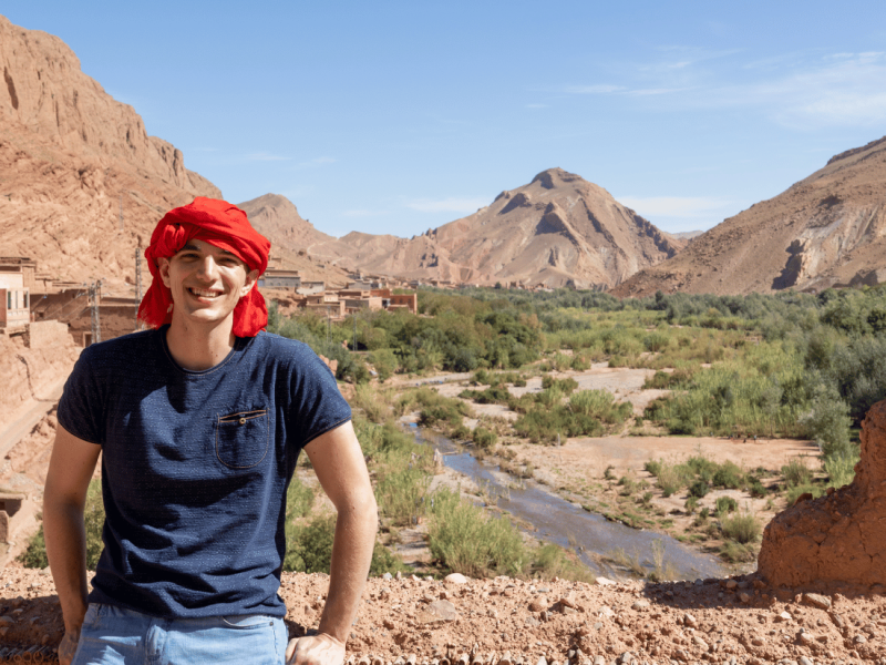 A traveler in a red headscarf leaning on an adobe wall with a Moroccan oasis landscape in the background