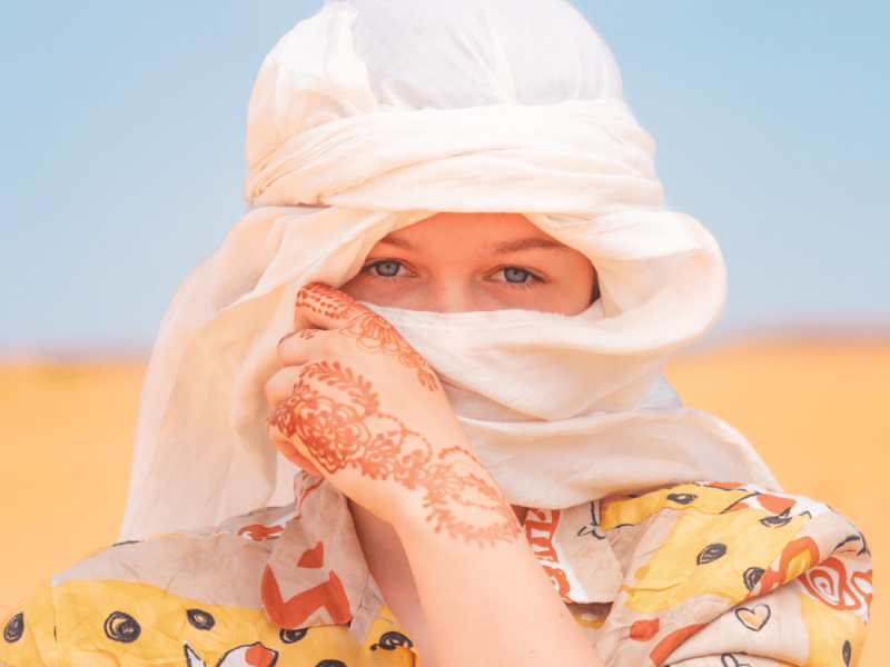A woman with blue eyes peering out from a white headscarf, adorned with henna patterns on her hand