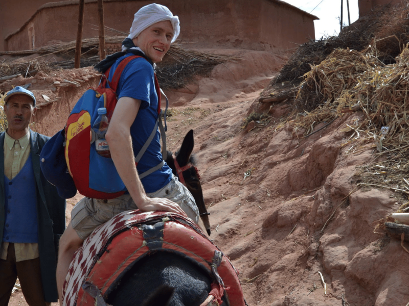 A traveler in a headscarf and backpack rides a mule, guided by a local, on a rural Moroccan trail.