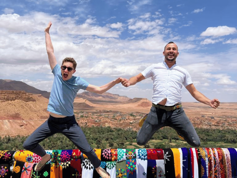 Two men happily jump above a vibrant collection of textiles, framed by a stunning Moroccan vista.