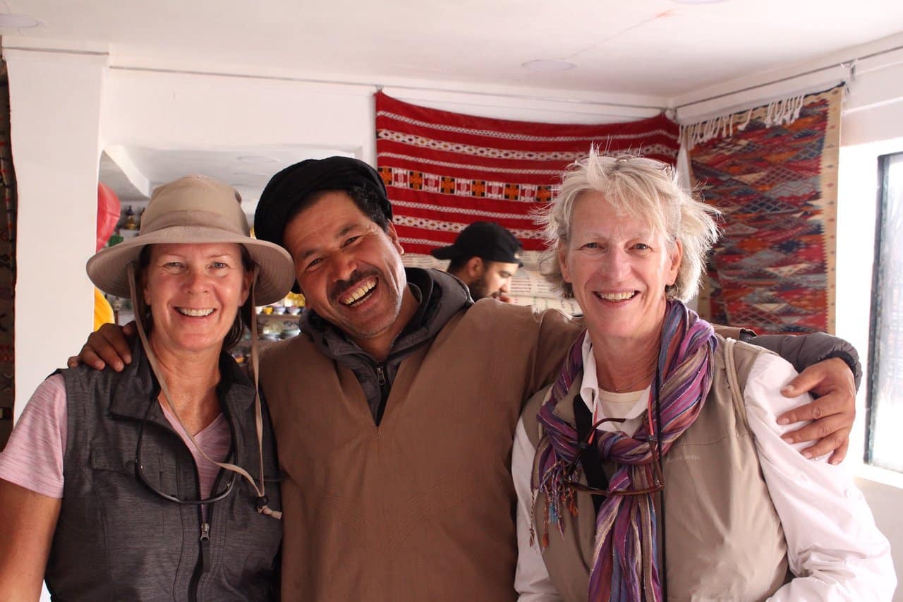 Two travelers smiling with a local Moroccan man inside a traditional rug shop. Travel to Morocco in January for warm hospitality and cultural experiences.