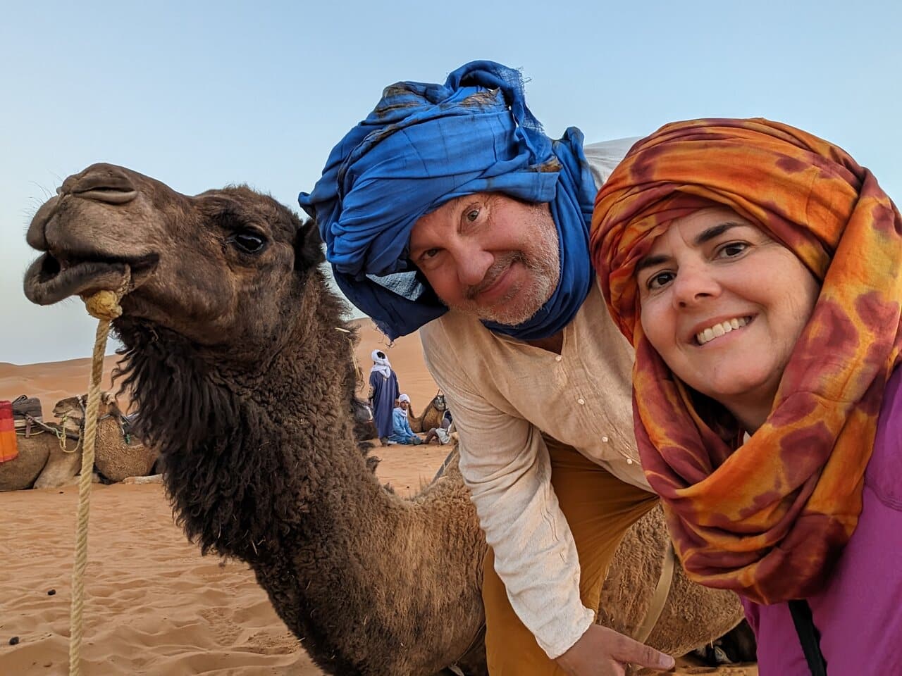 Two travelers wearing traditional scarves posing with a camel in the Moroccan desert. Visit Morocco in December for an unforgettable desert experience.