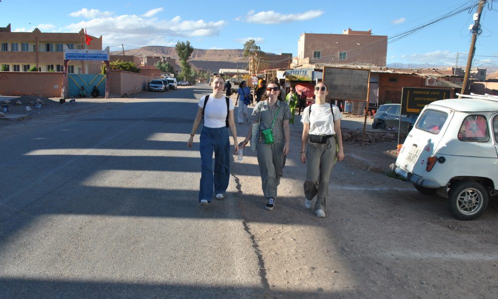 Three travelers walking along a village street in Morocco, surrounded by local shops, traditional buildings, and a vintage car. A cultural stop on An 8-Day Morocco Itinerary For Families.