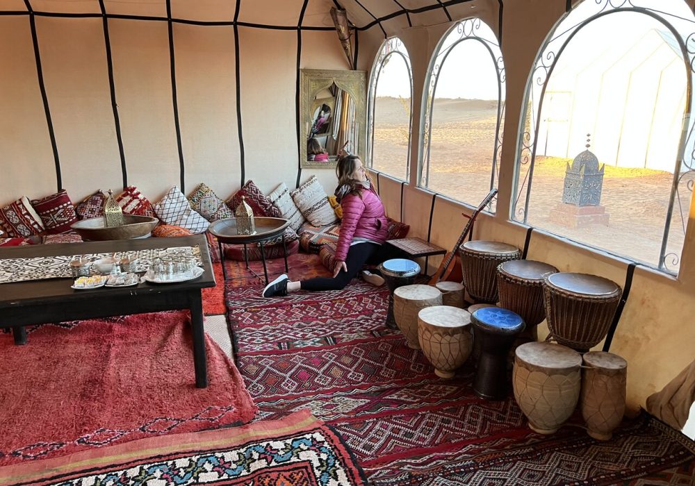 A traveler sitting inside a beautifully decorated desert camp lounge with traditional Moroccan carpets, cushions, and drums. Visit Morocco in December for an authentic desert experience.